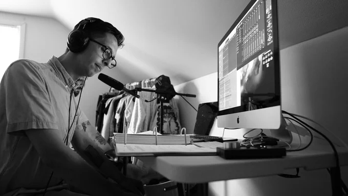 ID: A black and white photo of our sound designer Adam Raymonda sitting at his desk and reviewing the script during the production of season on.