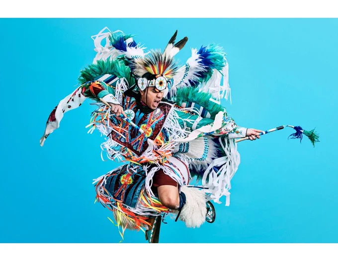 An image of the Notorious Cree, a men’s fancy Powwow dancer wearing brightly colored regalia in front of a bright blue background.