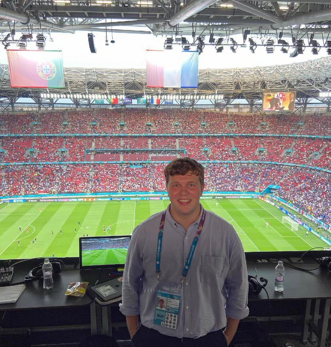 Brad. Audio description commentator working at euro 2020, stood with the stadium behind him in the commentary box. Players are on the pitch and the crowd are in the seats.
