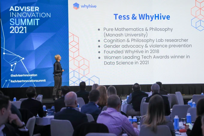 Young person with blonde hair, glasses and a black jacket on stage.   There is a projected presentation with a short bio reading “Tess and WhyHive, Pure Mathematics and Philosophy (Monash University), Cognition and Philosophy Lab researcher, Gender advocacy and violence prevention, Foundation WhyHive in 2018, Women Leading The Awards Winner in Data Science in 2021.”