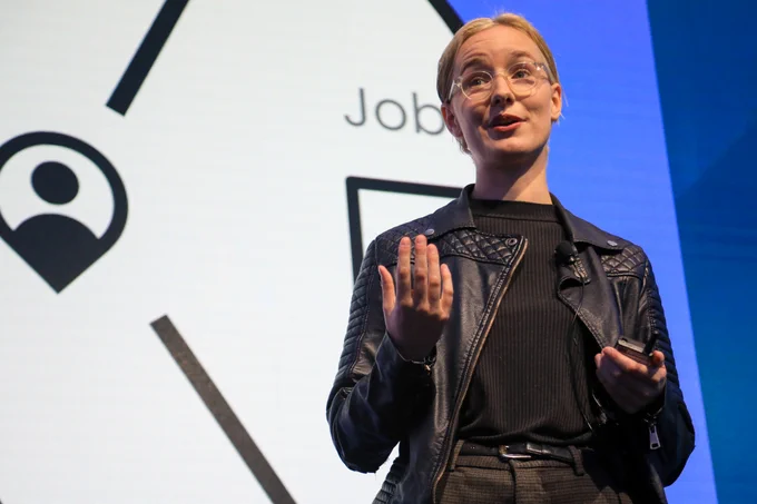 A close-up photo of a young person with blonde hair, glasses and a black jacket on stage giving a presentation. 