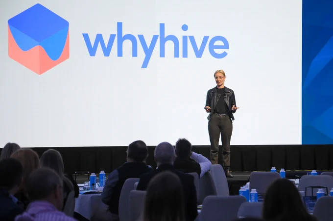 The WhyHive logo is projected behind them in the upper left. The logo is a red and blue hexagon, with whyhive written in light blue to text to the right.