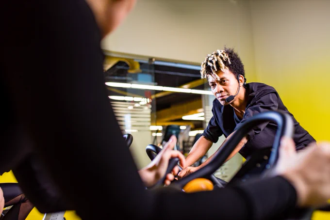Mixed race coach leads spin class session, encouraging group out of shot who are training on exercise bikes