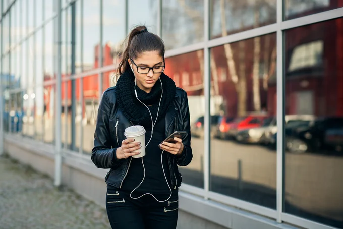 Person walking outside with headphones on holding a cup of coffee and a cell phone