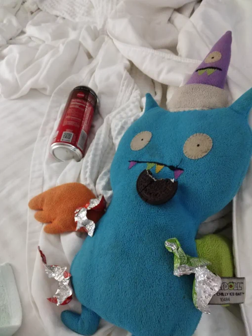 Photo of a Sleepy Chilly Ice Bat plush stuffed cartoon bat, the Gund Uglydoll, made of blue felt, with a green wing, an orange wing, and a purple hat which has its own eye and teeth. Ice Bat's teeth are multicolored. He is laying on a bed, with foil candy wrappers strewn on him, a half eaten oreo hanging out of his mouth, and laying next to him is a coca cola with coffee drink.