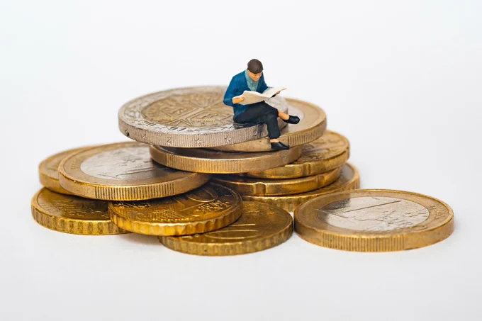 A pile of giant gold coins with a miniature figure sat on top of the pile reading a newspaper