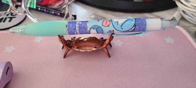 Photo is a close-up shot of a pink desk mat where a small, plastic, realistic-looking red little crab figurine holds up its claws. It is perfect for placing a pen in its little claws! It is indeed holding a blue pen and looks so proud of itself with its beady little plastic eyes.