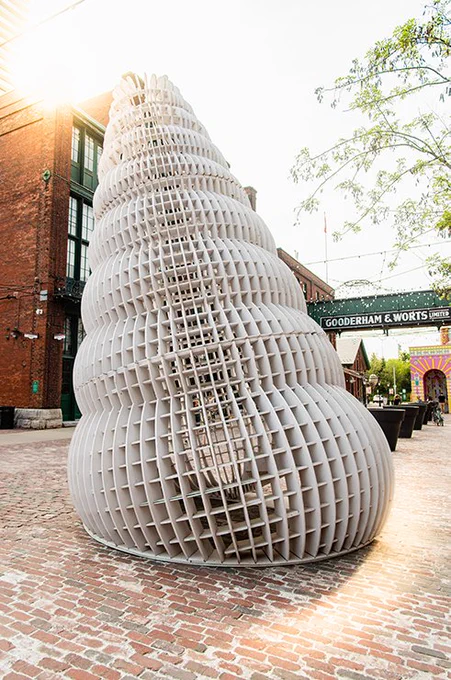 An image of The Epitonium installation. The structure is large, white, with a grid-like surface that allows the viewer to see through the installation. The installation is slightly tilted to one side, with the foundation of it being a large circular, bulbous shape. Moving up from that foundation, similar circular, bulbous shapes are stacked on top, but they are gradually smaller. In the background, we can see the Gooderham & Worts sign in the Distillery District, a brick building, and cobblestone lining the ground.