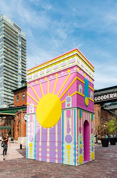 A photo of the Arc de Blob installation. The installation is large, rectangular, and multi-coloured. There is a yellow circle in the centre with yellow lines radiating from it. The background colour is mostly pink, and the surface is lined and decorated with shapes and motifs in purple, yellow, pink, blue, and light green. In the background, we see a condominium and bright blue sky behind the Distillery District buildings and cobblestone walkway.
