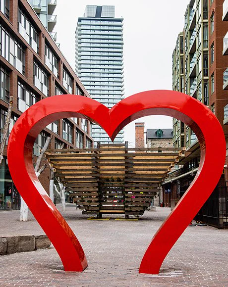 An image of the From Small Beginnings installation. The installation itself is out of focus in the background, with the focus being drawn to a heart shaped installation  in front of it that frames From Small Beginnings. In the background, From Small Beginnings consists of horizontal wooden boards, arranged together to allow a sliver of empty space in between. The boards on the bottom are shorter, and the ones above get gradually longer to form an upside down trapezoid shape. The ground is laid with cobblestone, and we see many buildings in the background of the Distillery District. 