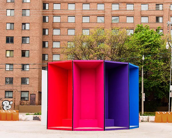 An image of the Throbber installation. The installation consists of multiple congruent parts of different colours that form a polyhedron, possibly an octagon, but it is unclear as we can only see 5 faces from this photo. The faces of the polyhedron are not flat, but concave to allow depth to the installation. The colours of the faces we can see are white, red, pink, purple and blue.  The background shows an apartment building with trees on the side of the road. 