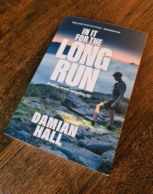 Photo shows the front cover of 'In It For The Long Run', with the title in large white letters over a picture of Damian at the top of a mountain. The peak is just above the cloud line, with a cloudy mist going off into the distance and only a couple of other peaks showing through.
