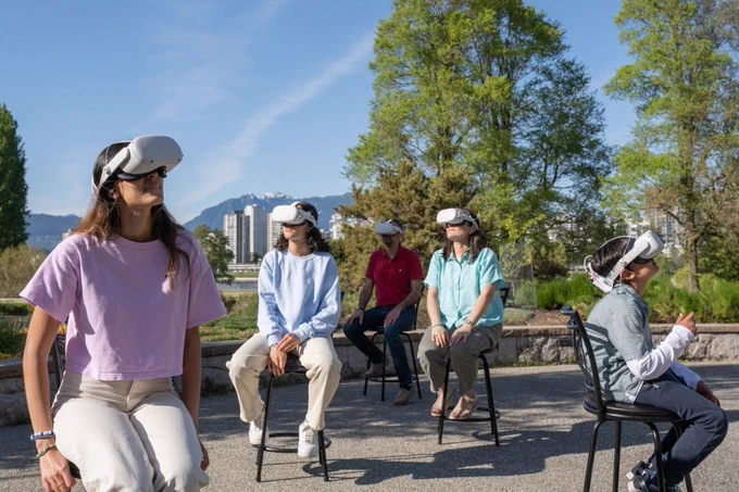 Five people wearing VR headsets sit on high stools outside. 