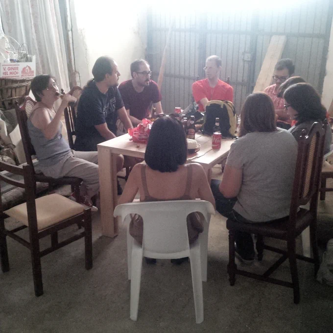 9 personas (4 mujeres, 5 hombres) alrededor de una mesa en la cochera de mi casa en el pueblo. Sobre la mesa, varios botellines de cerveza. Las sillas son una mezcla de lo que había por casa: sillas de plástico de terraza y sillas de madera antiguas.