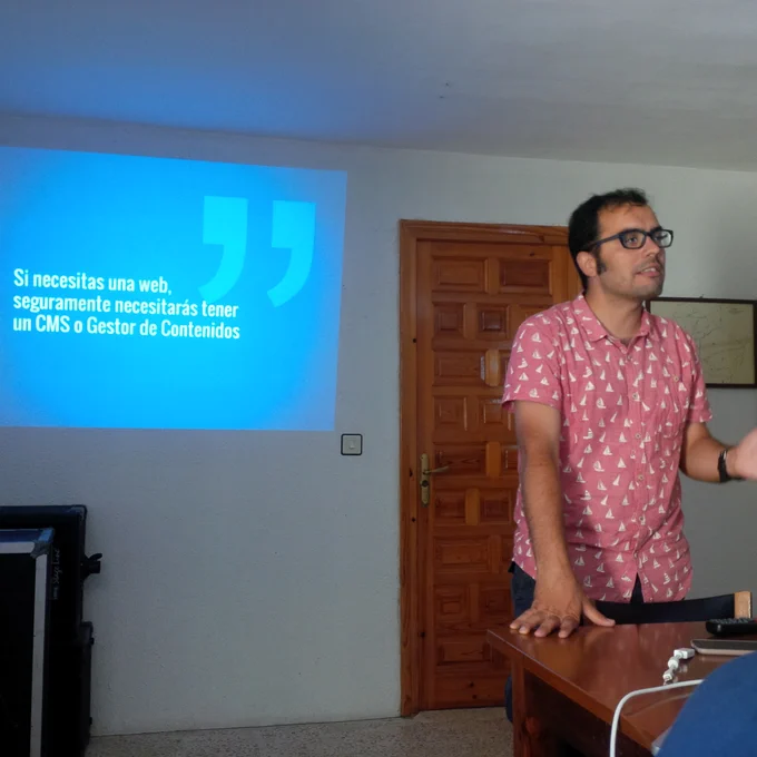 Un ponente (chico joven vestido de manera informal) hablando sobre Wordpress. Al fondo se ve la pantalla proyectada sobre la pared donde se lee: "Si necesitas una web, seguramente necesitarás tener un CMS o Gestor de Contenidos".