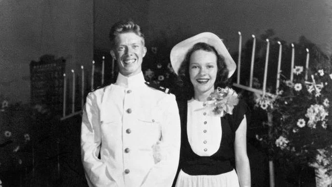 President Jimmy Carter and First Lady Rosalynn Carter on their wedding day on July 7, 1946. Jimmy is wearing is white Navy uniform while Rosalynn is wearing a light dress with dark sleeves and a hat. They stand in front of two candelabras.