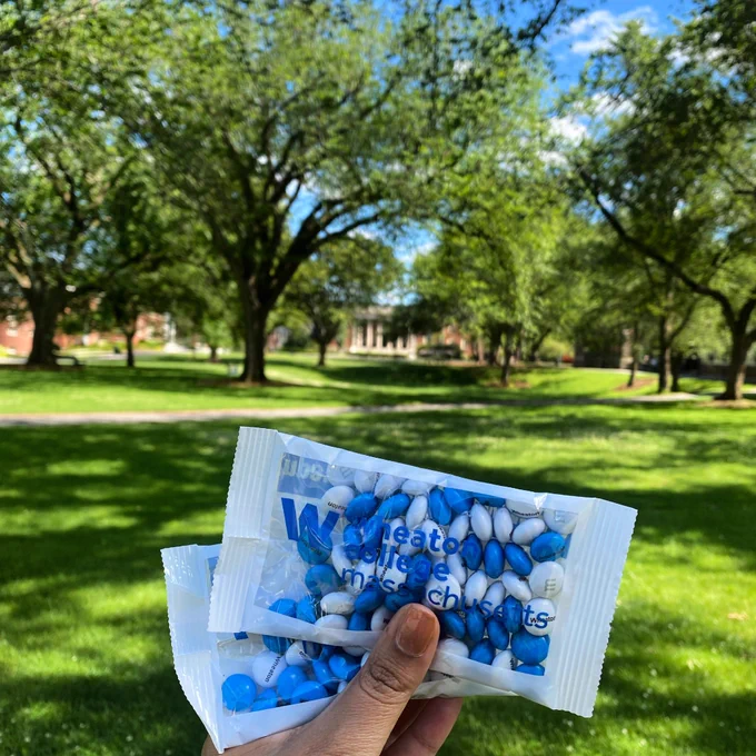 Hand holding two packs on blue and white M&M chocolates superimposed on a college quad