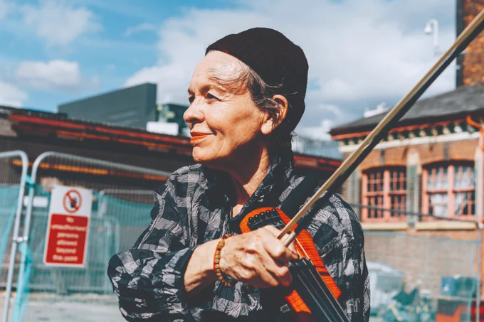  A chest-up portrait of Laurie Anderson holding her electric violin and standing outside in front of construction fencing, with a brick warehouse-style building behind. Laurie is white and 74 years old with short grey hair worn under a black beanie hat. She faces slightly to the left and looks far left with a closed-mouth smile. She is wearing coral lipstick, a black and grey checked shirt and a wooden beaded bracelet, and holds a long violin bow upward in her hand; her slender electric violin rests against her collarbone.