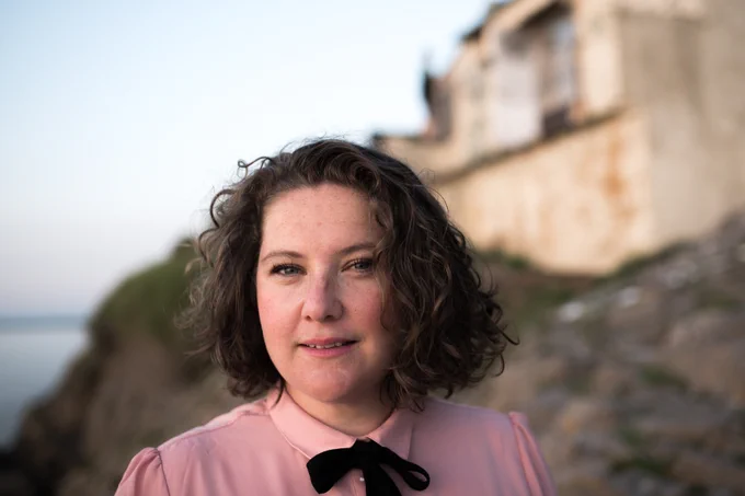 A headshot of Nuala, caucasian, soft curls loose in a bob, a dusty pink blouse with black bow tied at the collar in front of a blurred background of coastal house and cliff