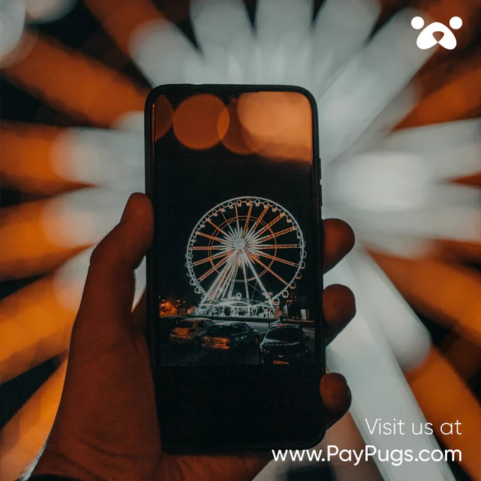An image of a hand holding a mobile phone while photographing an illuminated ferris wheel at night. The PayPugs image as well as the text ‘Visit  us at www.PayPugs.com’ at the bottom.