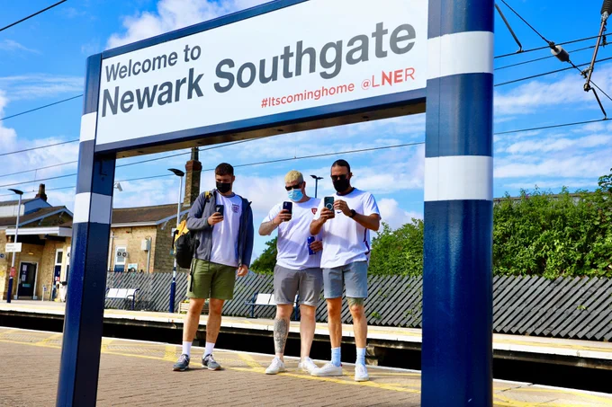 Three masked fans, and a sign that says 'Welcome to Newark Southgate #itscominghome @LNER'