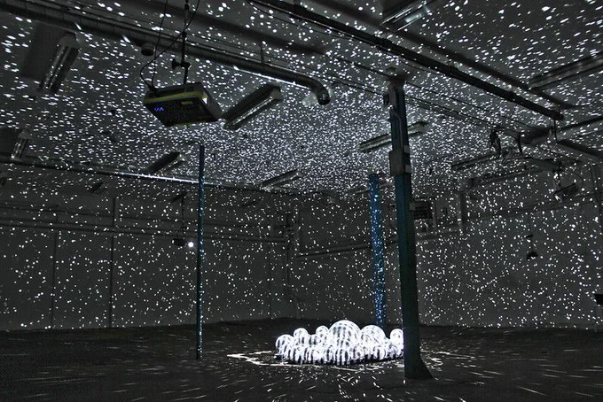 Image is installation view photograph of a dark room with thousands of pinpricks of light projected onto the walls, ceiling, and floor from a cluster of disco ball- like orbs installed among the support beams in the center of the room.