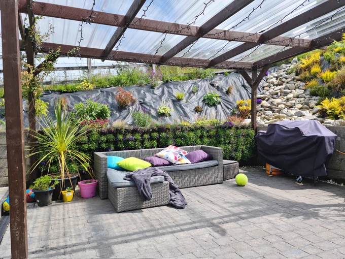 Outdoor sofa with colourful cushions under a pergola. Garden and behind. Bbq in the corner.