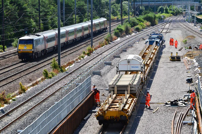 A track laying machine installs new sleepers and railway tracks while an LNER train heading to London speeds past on the existing track to the left.