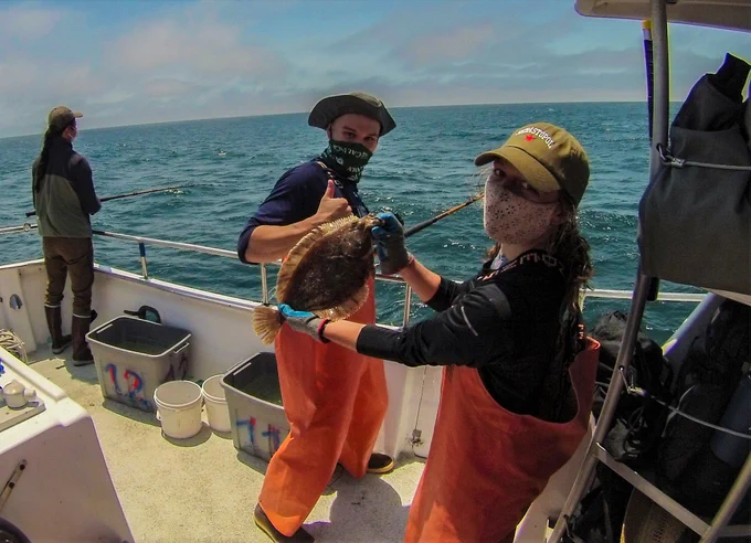 Science crew member holding a rock sole in front of angler on a boat. 