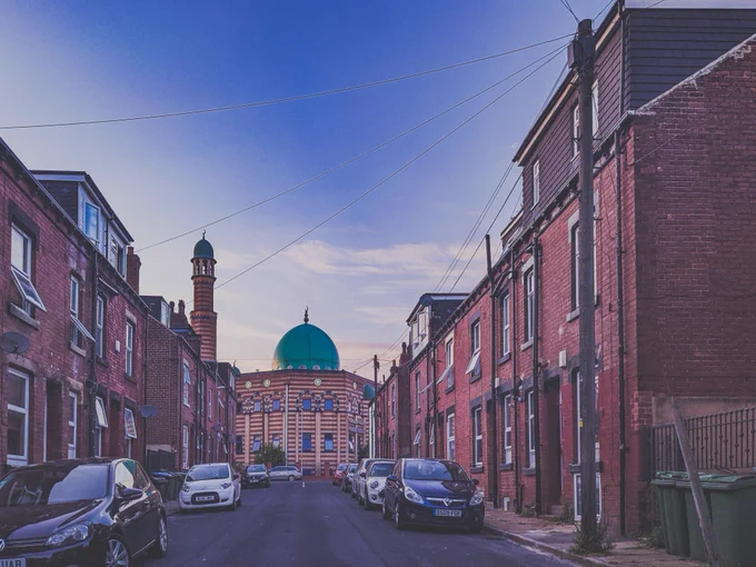 Mosque with a minaret and green dome is at the end of a street of terraced red brick houses. Cars line both sides of the streets.