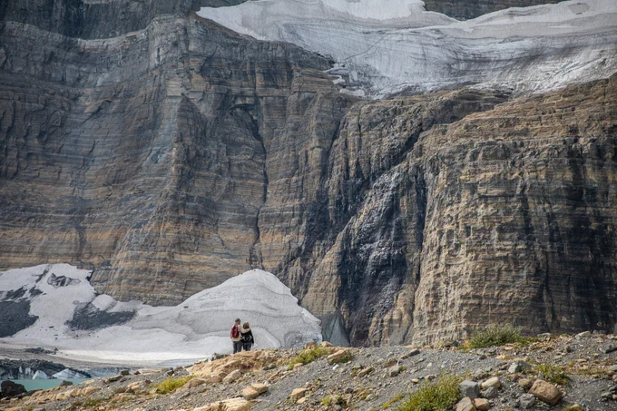 Two hikers stand on a rocky ridge in front of a glacially-carved amphitheater, with steep cliffs, two glaciers, and a blue glacial lake.