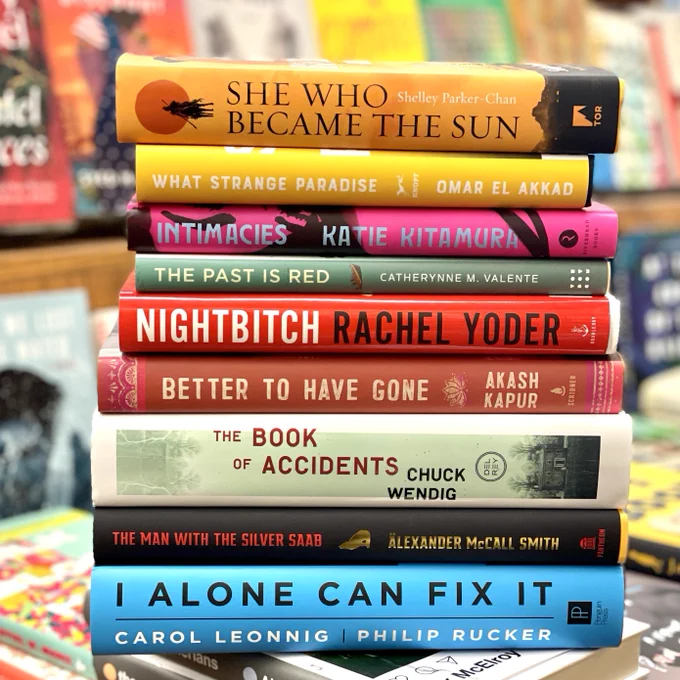 Stack of newly released hardcover books in a bookstore. She Who Became the Sun by Shelley Parker-Chan, What Strange Paradise by Omar El Akkad, Intimacies by Katie Kitamura, The Past is Red by Catherynne M. Valente, Nightbitch by Rachel Yoder, Better to Have Gone by Akash Kapur, The Book of Accidents by Chuck Wendig, The Man with the Silver Saab by Alexander McCall Smith, I alone Can Fix It by Carol Leonng and Philip Rucker