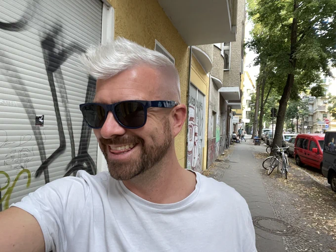 Selfie mit Sonnenbrille und silbernen Haaren. Im Hintergrund eine typische Straße in Berlin Neukölln mit Grafitis auf den Wänden.