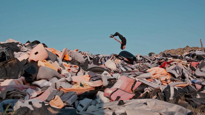 In this screenshot from The Story Won't Die, a person is seen in the distance doing a back flip over a mound of left behind backpacks that stretches the width of the image.