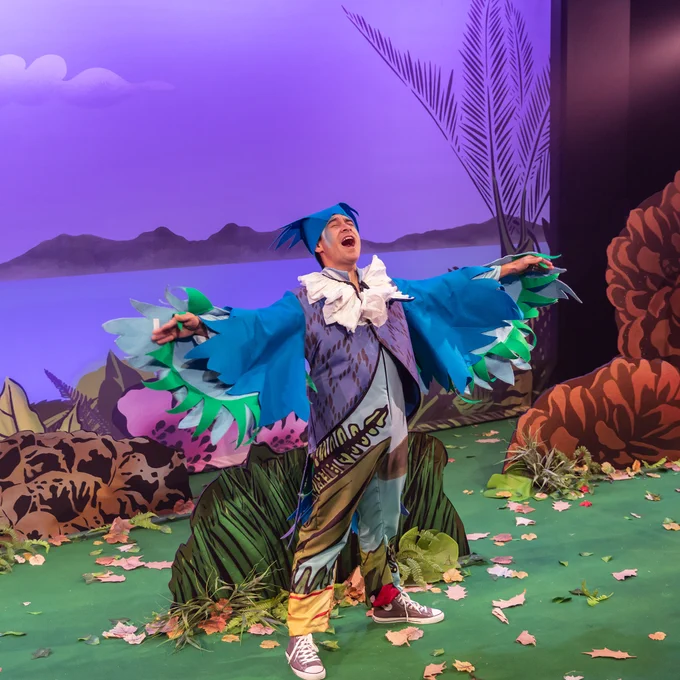 An actor on stage in a Tui bird costume stands with their arms (or wings!) outstretched.