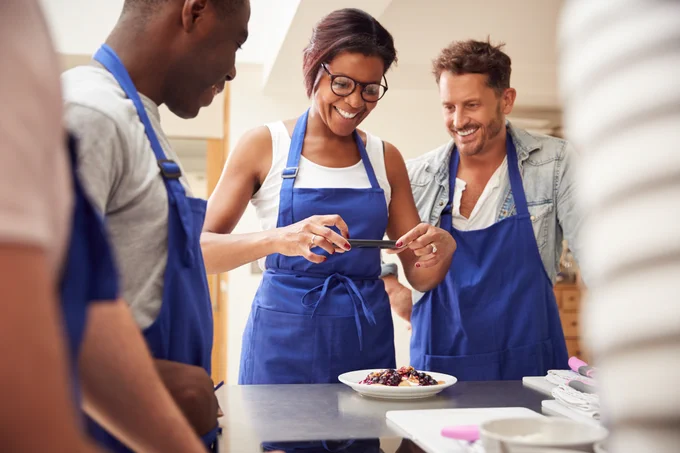Adult female student smiles as she takes a photo of pancakes with her phone. Two adult male classmates look on with smiles on their faces. They all wear blue aprons.