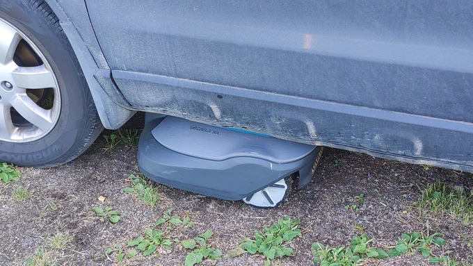 Robot lawnmower stuck under left side of car just behind the wheel, parallel with door.
