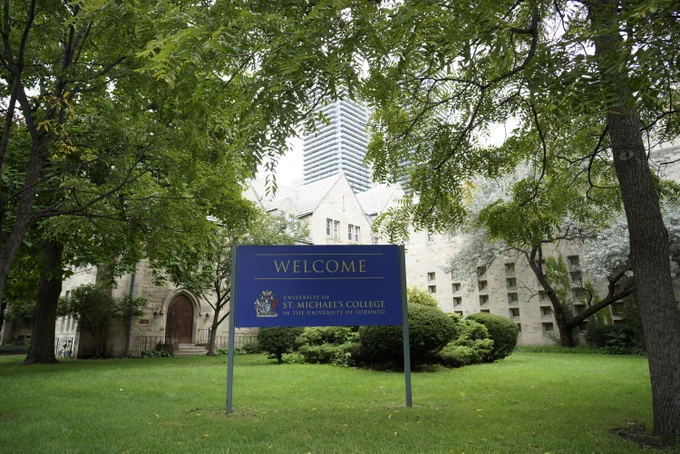 Photograph of the St. Mike's welcome sign with Carr Hall in the background and green trees surrounding