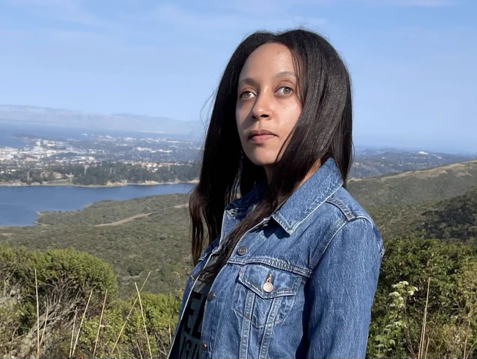 I’m standing on the side of a trail with green hills and a blue lake behind me. I’m wearing a denim jacket over a gray shirt, and my long black hair is framing my face.