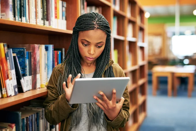 Student inside a library with a bookshelf behind her as she works on an iPad. 
