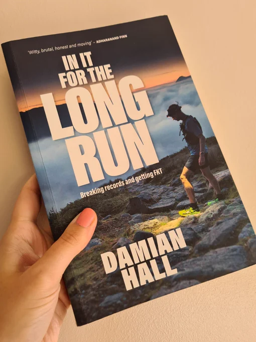 Photo shows front cover of In It For The Long Run, with the title in large text in front of a photo of Damian in running shorts and tshirt at the top of a hill. Behind him you can see the tops of the clouds that go out into the distance