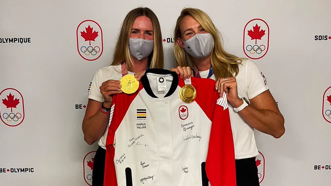 Two women with long blonde hair hold up a jacket signed by the women's eight crew. They are also holding up their gold medals with the jacket. Both women are wearing grey masks and white t-shirts.