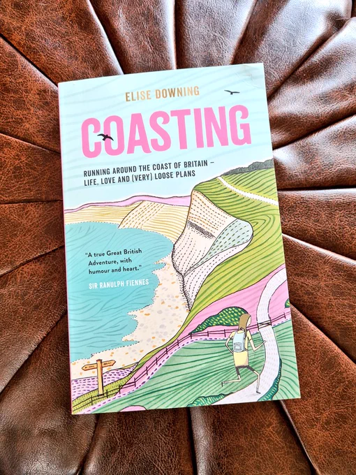 Photo shows the front cover of Coasting. It is an illustration of a path following along the edge of a cliff face, next to the sea. There is a small figure in the bottom corner running at the start of the path. The book sits on a brown footstool