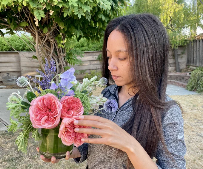 I’m holding up a bouquet with one hand, touching the flowers with the other, and leaning in slightly to breathe in the fragrances. The bouquet has lots of deep reds, purples, and greens. The photo was taken outside with lots of greenery in the background.
