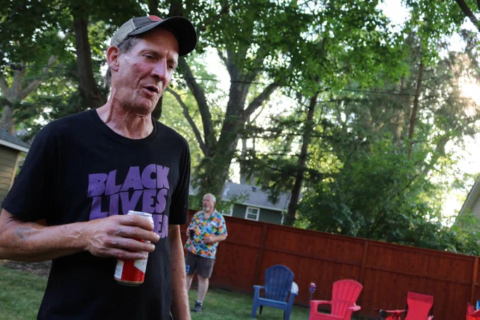 49 year old white guy who looks older than he is, holding a beer, wearing a baseball cap and a Black Lives Matter T-shirt in the style of Black Sabbath, standing in a suburban backyard