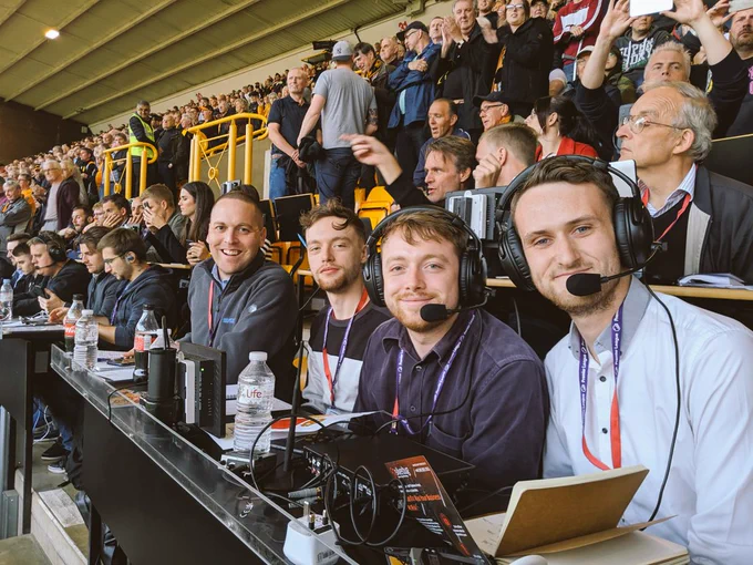 a team of 4 commentators in the press box with earphones and commentary equipment. The ground is ful of fans behind them. 