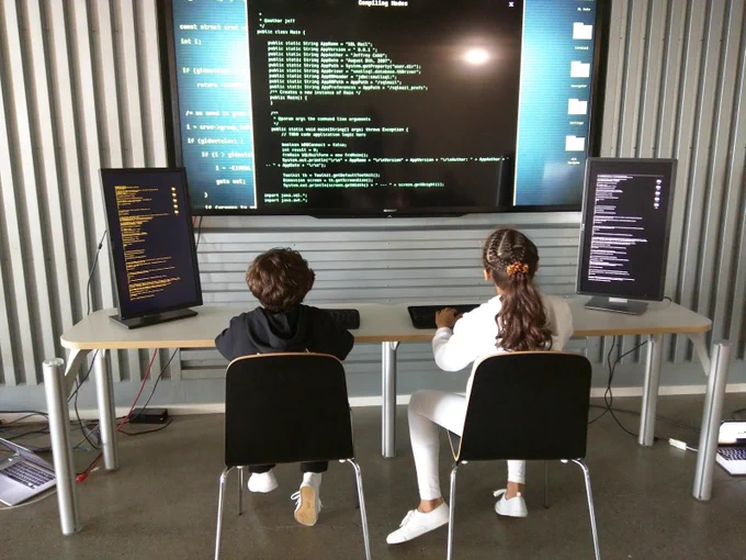 two kids sitting in front of huge monitor with code on it