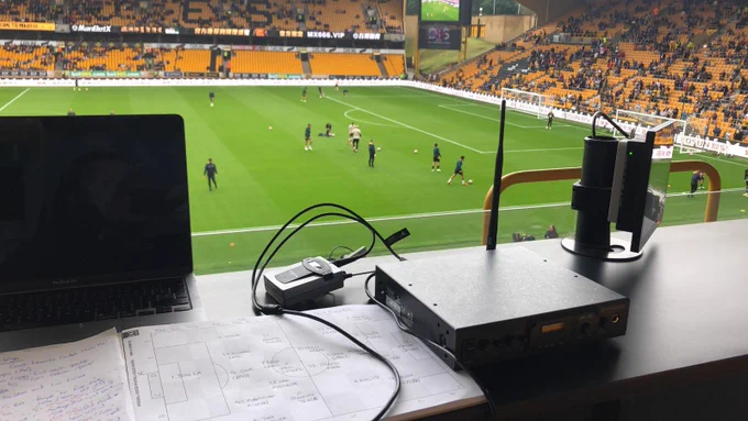 Press box at wolves with the pitch in the back ground with some players warming up. The stands half full abs AD equipment is on a desk ready to go