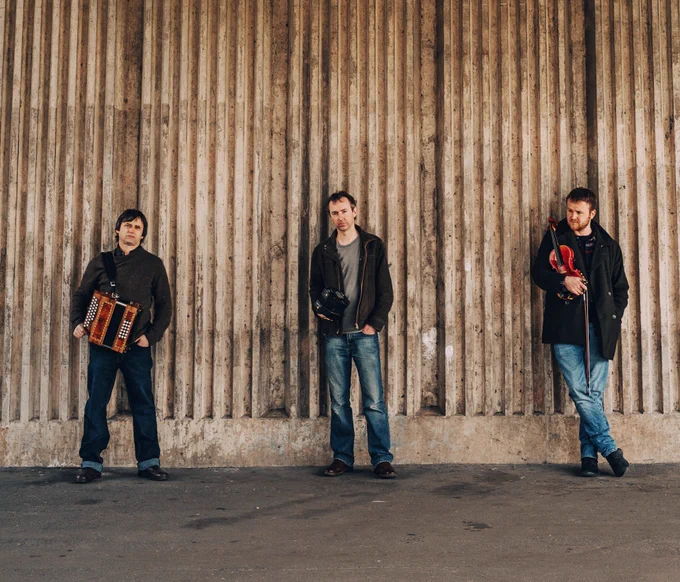 Andy Cutting with a melodeon, Rob Harbron with a concertina and Sam Sweeney with a violin, stood in front of a wall. 