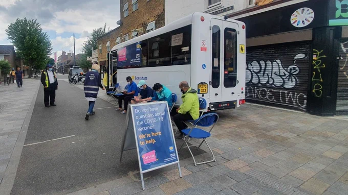 People are sitting on chairs outside a parked bus. They are wearing masks and filling out forms. There is a sign up reading 'Do you have questions about the COVID-19 vaccine? We're here to help.'